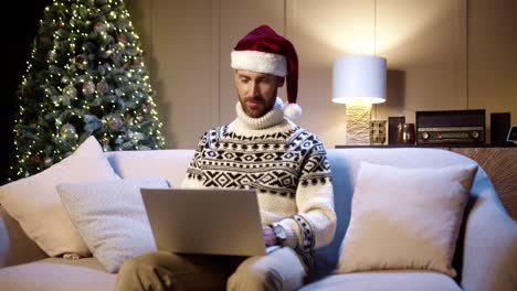 Portrait-Of-Happy-Young-Man-Wearing-Santa-Hat-Sitting-In-Decorated-Room-Near-Glowing-Christmas-Tree-Tapping-And-Browsing-On-Laptop