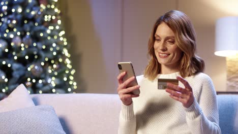 Primer-Plano-Retrato-De-Mujer-Hermosa-Positiva-Feliz-Comprando-En-Internet-Regalos-De-Navidad-Pagando-Con-Tarjeta-De-Crédito-Usando-El-Teléfono-Celular-En-Una-Acogedora-Casa-Decorada