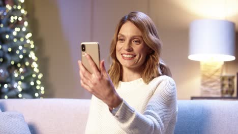 Close-Up-Portrait-Of-Joyful-Woman-Speaking-On-Video-Call-Online-On-Smartphone-Receiving-Holidays-Greetings-On-Christmas-Eve-While-Sitting-In-Cozy-Room-Near-Glowing-Xmas-Tree-New-Year-Concept