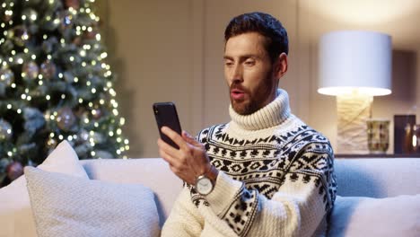 Nahaufnahme-Eines-Glücklichen-Jungen-Mannes-Mit-Weihnachtspullover,-Der-In-Einem-Zimmer-In-Der-Nähe-Eines-Geschmückten-Leuchtenden-Weihnachtsbaums-Sitzt-Und-Online-Auf-Dem-Handy-Chattet
