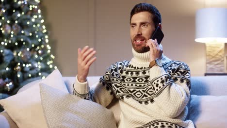 Close-Up-Portrait-Of-Joyful-Young-Man-Wearing-Xmas-Sweater-Sitting-In-Room-Near-Decorated-Glowing-Christmas-Tree-Speaking-On-Cellphone-Telling-Holiday-Greetings