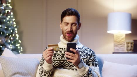 Close-Up-Portrait-Of-Cheerful-Young-Man-Makes-Purchase-On-Internet-Buying-Xmas-Gifts-On-Cellphone-Paying-With-Credit-Card-At-Home-Near-Glowing-Tree