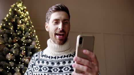 Close-Up-Portrait-Of-Happy-Male-In-Good-Mood-Videochatting-On-Cellphone-Near-Glowing-Decorated-Xmas-Tree