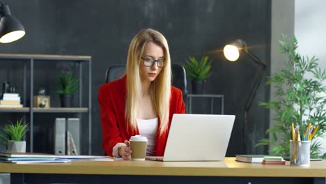 Young-Blonde-Woman-Working-Sitting-At-Desk-In-Front-Of-Computer-In-The-Office,-Then-She-Drinks-Coffee-To-Go