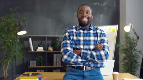 Handsome-Man-In-Plaid-Shirt-Sitting-On-Table-In-Office-Room,-Crossing-Hands-And-Smiling-To-Camera
