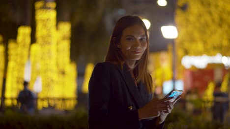 Atrractive-young-woman-checks-her-smartphone-then-smiles-at-the-camera-under-the-bright-city-lights---blurred-background