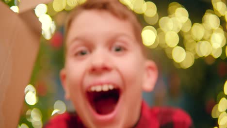 Fröhlicher-Junge-Vor-Weihnachtsbaum-Bricht-Durch-Karton-Und-Zeigt-Sein-Breites-Lächeln