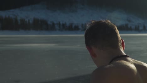 Young-man-with-short-hair-and-mustache-sitting-shirtless-beside-frozen-mountain-lake-with-snow-on-the-ground