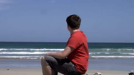 Male-in-contemplation-sitting-on-beach-rock-shore,-looking-out-to-ocean-waves-seascape