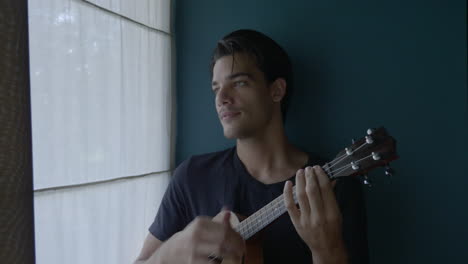Happy-Young-Male-Playing-Ukelele-In-Front-Of-Window,-Soft-Light
