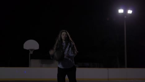 Teenage-girl-with-long-brown-hair-dribbles-ball,-throws-it-up-and-catches-it-before-shooting-at-a-dark-outdoor-court-with-lights