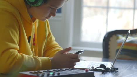 Teenager-Auf-Einem-IPhone-Und-Kopfhörer,-Während-Er-An-Einem-Laptop-Mit-Midi-Tastatur-Am-Tisch-Sitzt