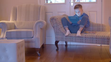 Young-child-playing-games-on-a-tablet-sitting-alone-on-a-chaise-longue-at-home-during-the-day