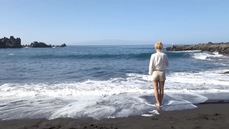 Lonely-Woman-Standing-on-Beach-While-Ocean-Waves-Breaking-on-Black-Sand-Coast-of-Tenerife,-Canary-Island,-Spain