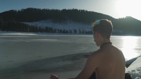 Young-man-with-no-shirt-sitting-beside-frozen-mountain-lake-in-winter-with-snow-on-landscape-and-bright-sunlight