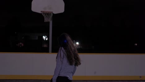 Teenage-girl-with-black-hair-bounces-ball-then-shoots-it-in-at-outdoor-court-with-grey-sweater-at-night