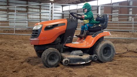 Two-Year-Old-Toddler-in-Dino-Costume-and-Cowboy-Boots-Sitting-Playing-on-Riding-Lawn-Mower-Inside-Arena