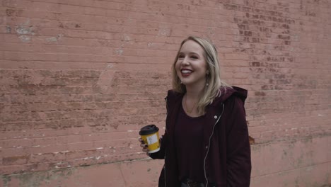Young-Blonde-Female-Smiling-and-Walking-Down-the-Street-with-a-Coffee-in-Hand-During-the-Day