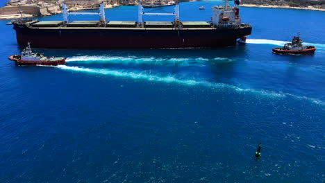 A-steady-aerial-profile-shot-of-a-giant-cargo-ship-leaving-the-harbour-of-Valletta,-Malta-with-the-help-of-three-tugboats