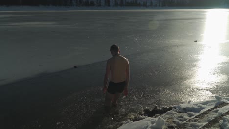 Bright-sunlight-shines-on-frozen-surface-of-mountain-lake-as-man-with-only-shorts-walks-into-the-water-where-ice-has-been-broken