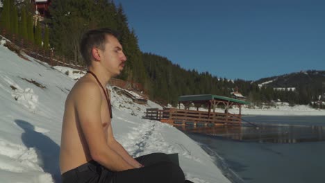 Young-man-with-no-shirt-sitting-in-snow-beside-frozen-mountain-lake-breathing-heavily