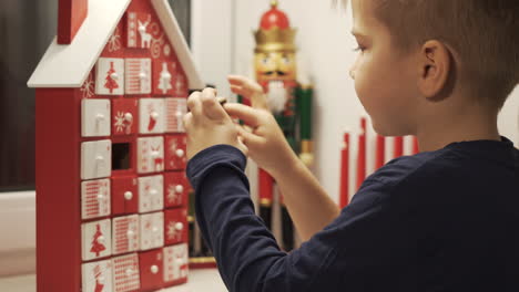 Glücklicher-Kleiner-Junge-Nimmt-Süß-Aus-Adventskalenderhausform-Mit-Weihnachtsnussknackern-Im-Hintergrund