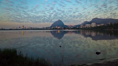 Zeitraffer-Eines-Sonnenaufgangs-Am-Stadtsee-Rodrigo-De-Freitas-In-Rio-De-Janeiro-Mit-Dem-Ufer-Im-Vordergrund-Und-Vorbeiziehenden-Wolken