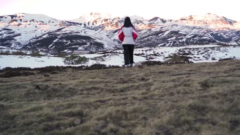 Slow-motion-shot-of-a-woman-dressed-in-warm-clothes-walking-across-a-grassy-field-looking-at-snow-capped-mountains