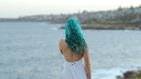 Leaving,-barefoot-blue-haired-young-woman-in-white-flowing-dress-turns-and-walks-towards-ocean,-copy-space