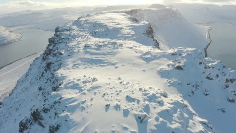 AERIAL-BIRDSEYE-TILT-DOWN:-Tracking-along-peak-ridge-of-Icelandic-mountain-range,-deep-snow-jagged-rocks-beautiful-sun-set