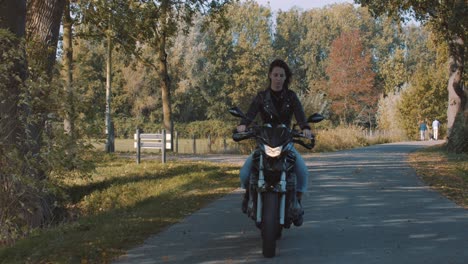 Pretty-smiling-European-young-woman-driving-a-motorbike-wearing-leather-jacket-in-forest-with-vibrant,-colorful-golden-autumn-leaves-on-sunny-day-1