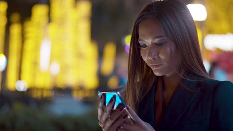 Attractive-young-woman-checking-her-smartphone-in-the-lights-of-the-city-at-night---background-bokeh-blur