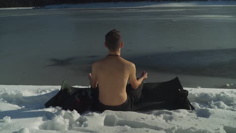 Man-with-no-shirt-sitting-on-black-yoga-mat-in-snow-beside-frozen-lake-meditating