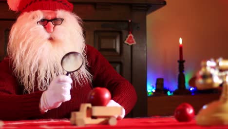 Santa-Claus-With-Magnifying-Glass-Inspects-Apples-On-A-Wooden-Toy-Truck