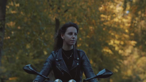 Pretty-smiling-European-young-woman-driving-a-motorbike-wearing-leather-jacket-in-forest-with-vibrant,-colorful-golden-autumn-leaves-on-sunny-day