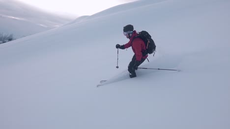 Zeitlupenaufnahme-Eines-Mannes-Mit-Roter-Jacke,-Der-Bergab-Ski-Fährt,-Umgeben-Von-Schneeweißen-Bergen-Und-Halbsonnigem-Hintergrund,-Der-Mit-Wolken-Bedeckt-Ist