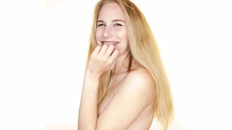 In-slow-motion,-against-a-white-backdrop,-a-young-blonde-woman-looks-over-her-bare-shoulders-and-shrugs-flirtatiously