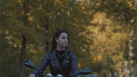 Pretty-smiling-European-young-woman-driving-a-motorbike-wearing-leather-jacket-in-forest-with-vibrant,-colorful-golden-autumn-leaves-on-sunny-day-4