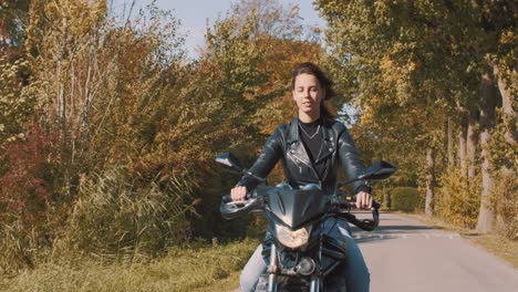 Pretty-smiling-European-young-woman-driving-a-motorbike-wearing-leather-jacket-in-forest-with-vibrant,-colorful-golden-autumn-leaves-on-sunny-day-3