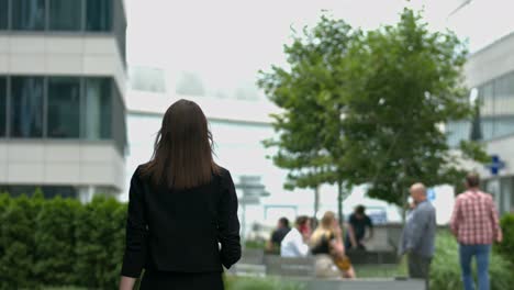 A-woman-with-long-brown-hair-walks-away-from-the-camera,-in-a-modern-urban-setting