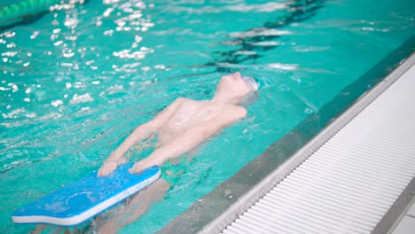 Boy-is-doing-backstrokes-in-indoor-swimming-pool-while-holding-a-paddle-board