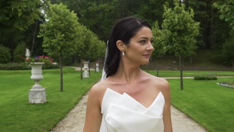 Front-view-of-the-beautiful-bride-walking-with-a-smile-confidently-in-the-park-on-her-wedding-day,-holding-her-wedding-dress-slit-and-showing-sexy-legs,-the-camera-follows-in-the-summer-day