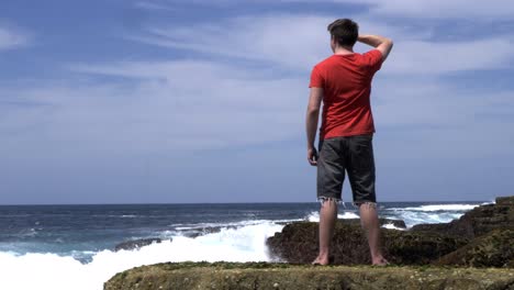 Male-standing-on-coastal-rock-beach-shore-watching-ocean-waves-in-contemplation,-meditation