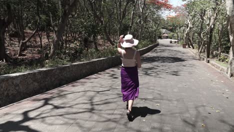 Young-female-tourist-with-a-white-hat-and-long-violet-skirt-walking-into-the-park,-Indonesia,-camera-follows-and-parallax