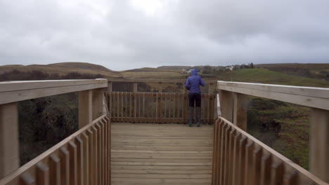 Tracking-follow-shot-of-happy-girl-walking-on-the-wooden-bridge-near-the-edge-of-a-cliff-with-hills-in-the-background-on-a-cloudy-day-in-Scotland,-Isle-of-Skye