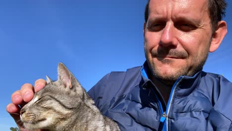 Closeup-shot-of-smiling-adult-man-cuddling-cat-outside-in-the-sunshine-at-blue-sky
