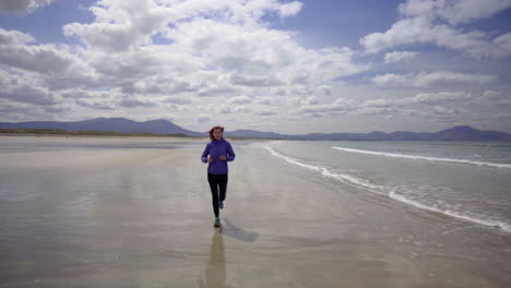 Dolly-shot-of-a-girl-running,-jogging-on-the-shore-of-a-sandy-beach-with-Atlantic-ocean-waves-on-a-wonderful-sunny-day-in-Ireland-in-4K