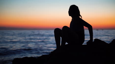 Eine-Junge-Dame,-Die-Auf-Den-Felsen-An-Der-Meeresküste-Sitzt,-Während-Sie-Ihre-Tägliche-Morgengymnastik-Macht,-Mit-Einem-Schönen-Sonnenaufgang-Im-Hintergrund---Weitschuss