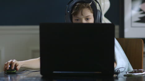 Medium-shot-of-young-boy-working-on-a-laptop-with-headphones-on-at-the-dining-room-table