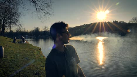 Young-man-with-glasses-standing-beside-a-lake-at-sunset-smoking-a-cigarette-exhales-blowing-a-stream-of-smoke-from-his-mouth